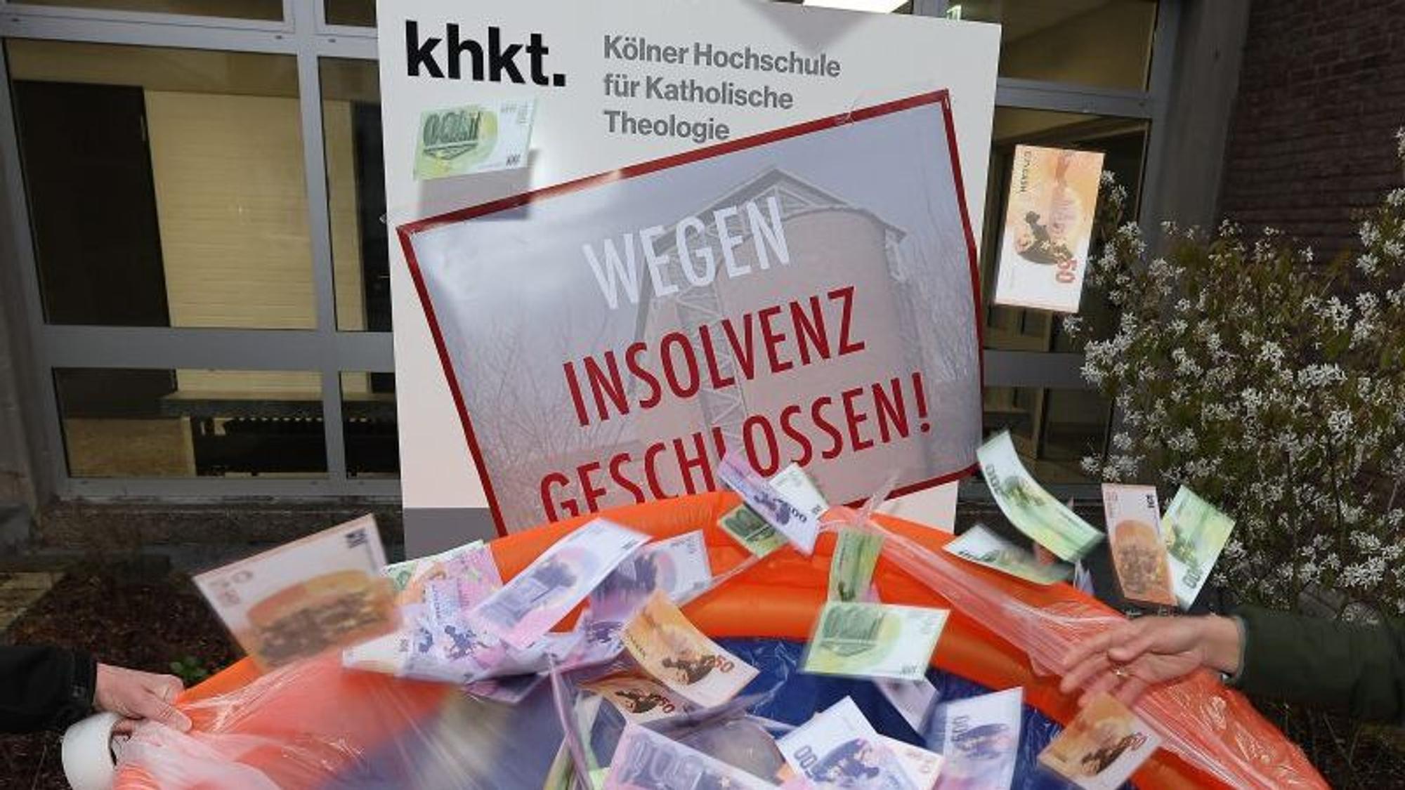 Geldscheine fallen in ein Planschbecken vor der Kölner Hochschule für Katholische Theologie (KHKT). Das Türschild am Eingang ist mit einem Plakat überklebt, auf dem „wegen Insolvenz geschlossen“ steht. Mit dieser Aktion wollten Kritiker der KHKT auf den hohen Finanzbedarf der Einrichtung aufmerksam machen.