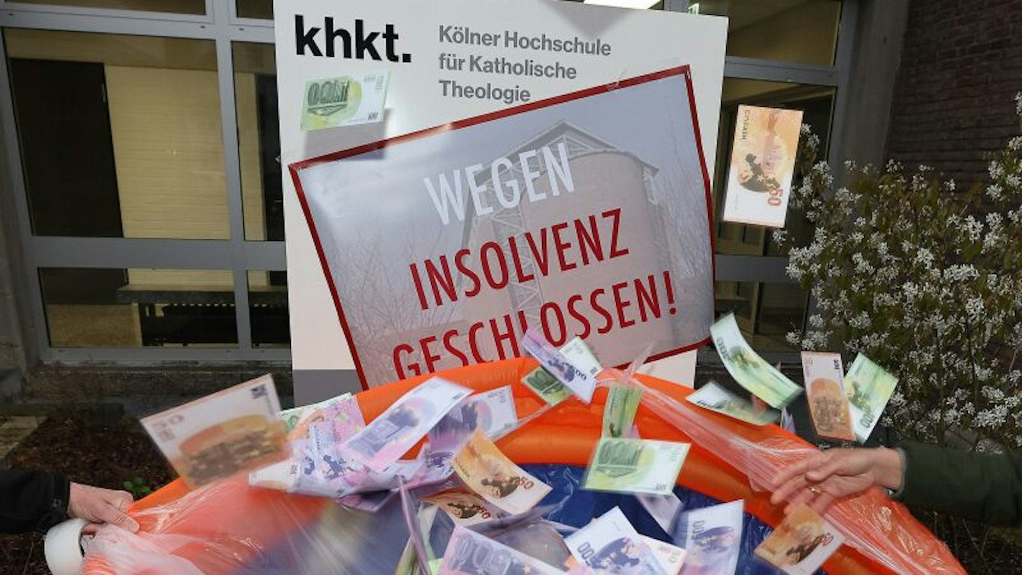 Geldscheine fallen in ein Planschbecken vor der Kölner Hochschule für Katholische Theologie (KHKT). Das Türschild am Eingang ist mit einem Plakat überklebt, auf dem „wegen Insolvenz geschlossen“ steht. Mit dieser Aktion wollten Kritiker der KHKT auf den hohen Finanzbedarf der Einrichtung aufmerksam machen.