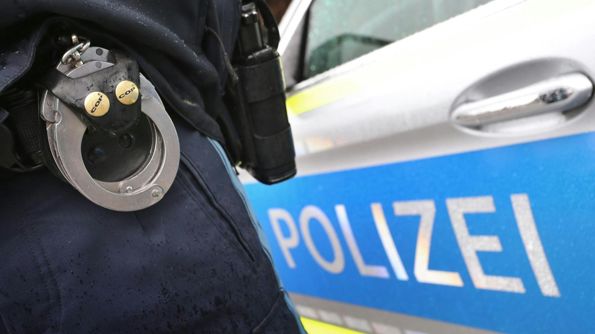 Auf dem Bild ist ein Polizeiauto und ein Polizist mit Handschellen zu sehen.