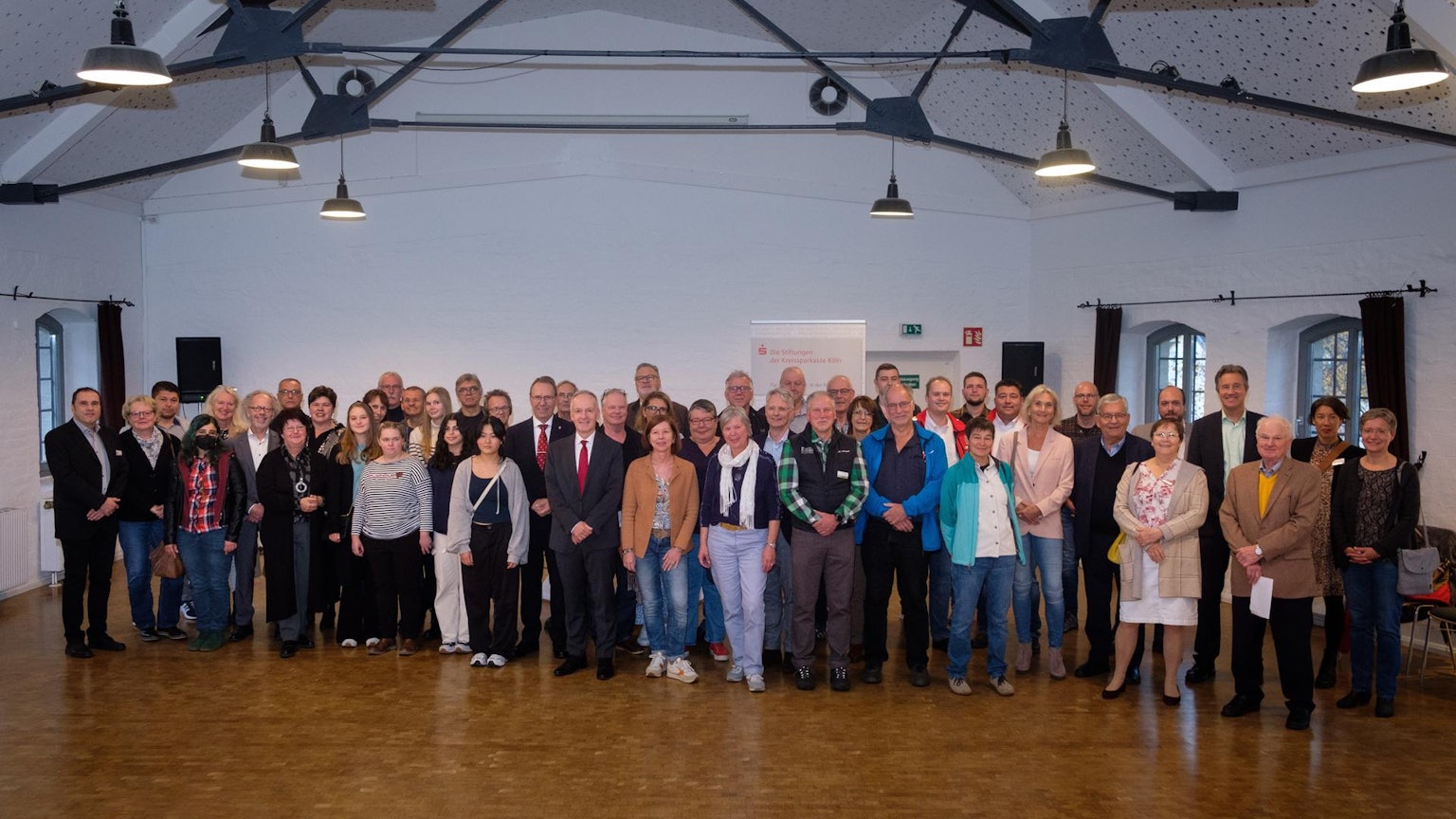 Die Mitglieder der teilnehmenden Stiftungen posieren zusammen vor einem Plakat der Sparkasse.
