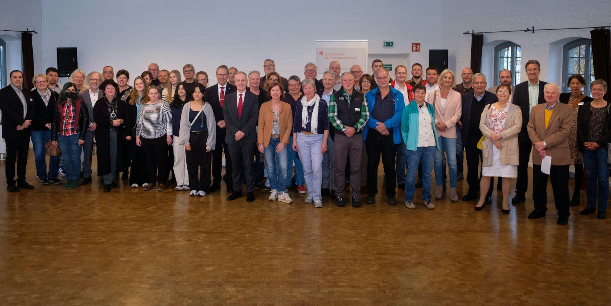 Die Mitglieder der teilnehmenden Stiftungen posieren zusammen vor einem Plakat der Sparkasse.