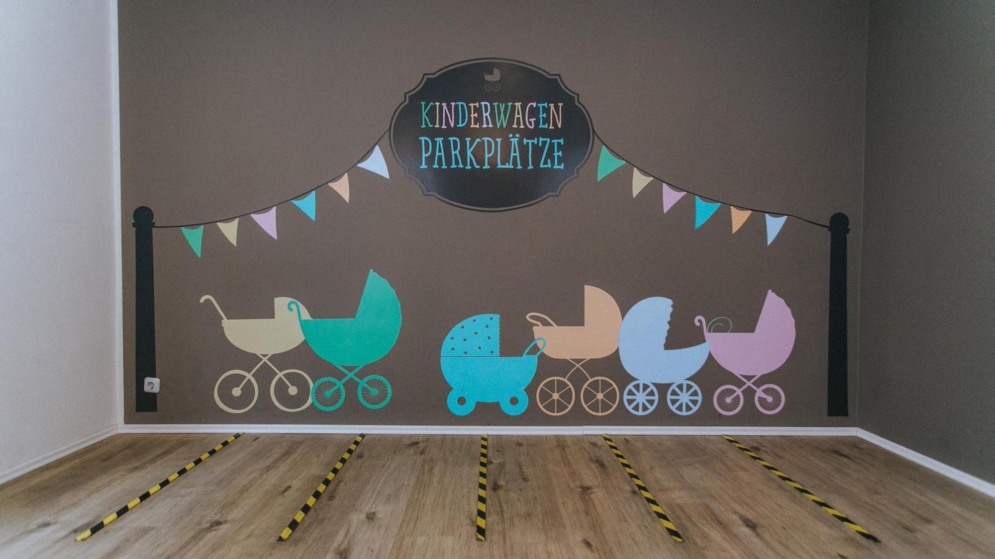 Kinderwagen auf eine Wand gemalt, darüber steht „Kinderwagen-Parkplätze", auf dem Boden sind rechteckige Felder markiert