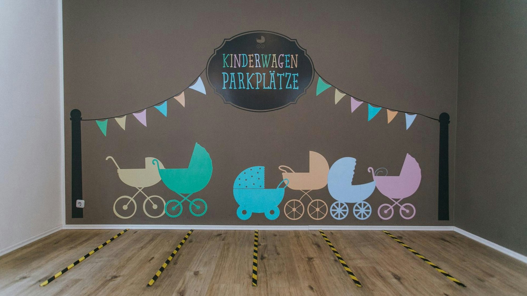 Kinderwagen auf eine Wand gemalt, darüber steht „Kinderwagen-Parkplätze", auf dem Boden sind rechteckige Felder markiert