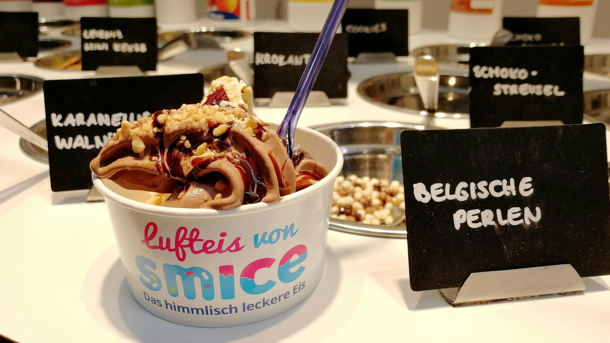 Ein Frozen-Yogurt-Becher von smice steht auf der Theke. Im Hintergrund sind verschiedene Toppings zu sehen.