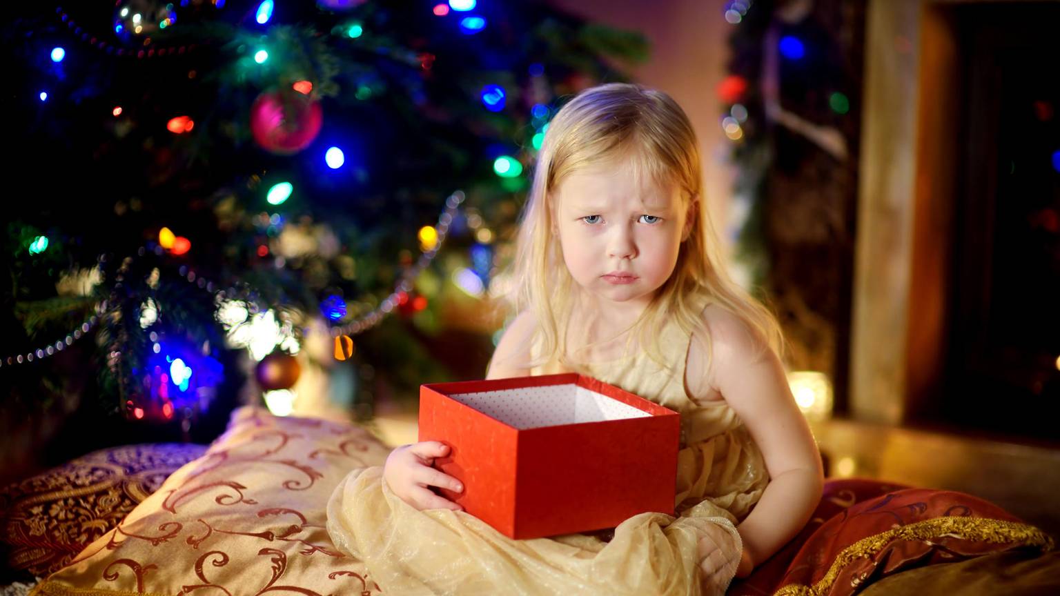 Ein Kind hält ein Weihnachtsgeschenk und sitzt vor dem Tannenbaum, es schaut schlecht gelaunt.