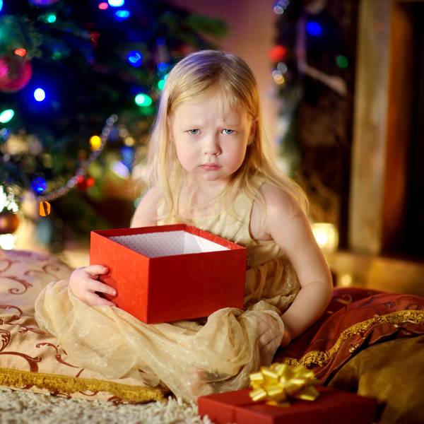 Ein Kind hält ein Weihnachtsgeschenk und sitzt vor dem Tannenbaum, es schaut schlecht gelaunt.