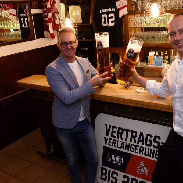 Gaffel-Chef Heinrich Philipp Becker steht mit FC-Geschäftsführer Alexander Wehrle.vor einer Theke. Sie stoßen mit übergroßen Kölsch-Gläsern an.