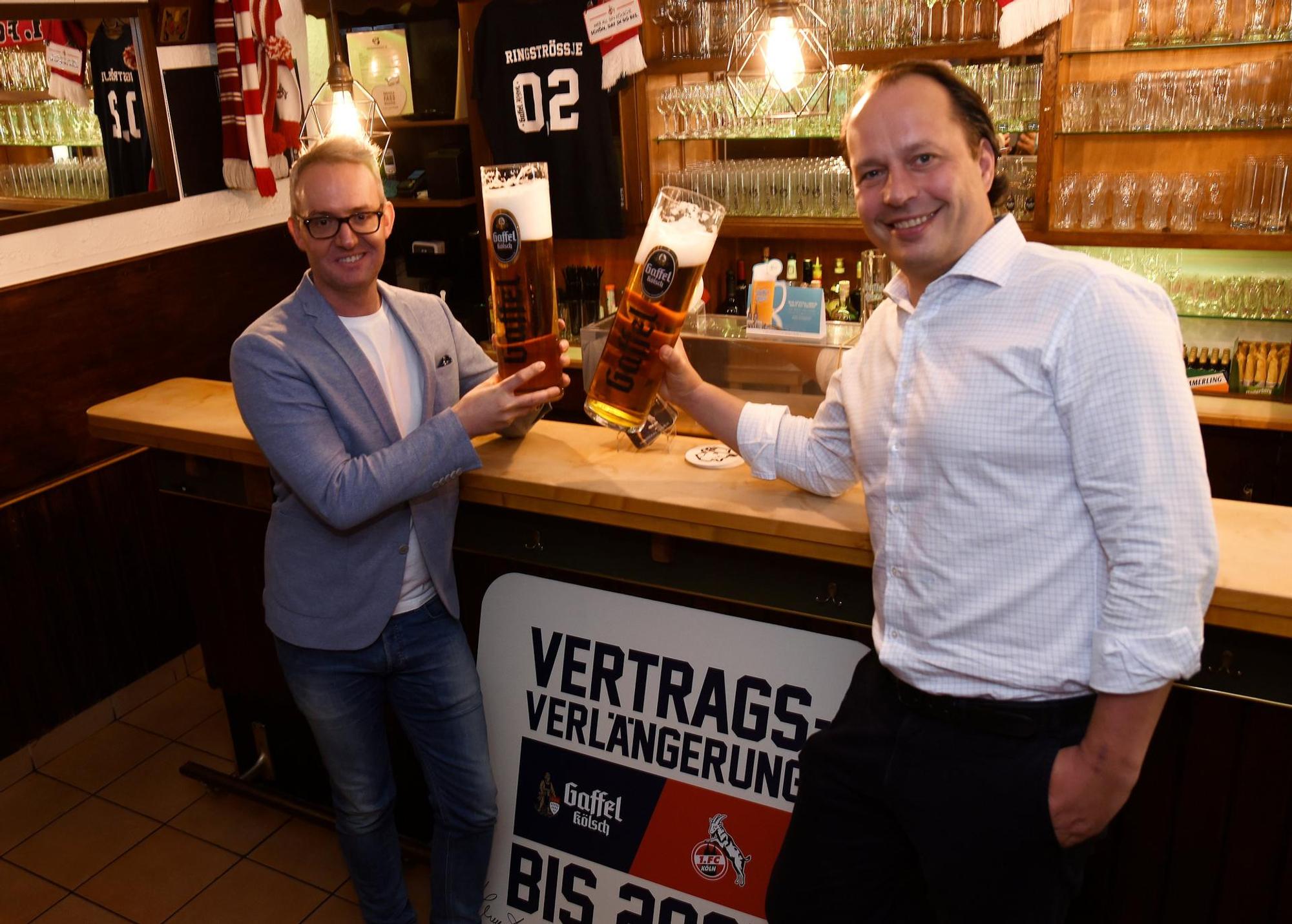 Gaffel-Chef Heinrich Philipp Becker steht mit FC-Geschäftsführer Alexander Wehrle.vor einer Theke. Sie stoßen mit übergroßen Kölsch-Gläsern an.