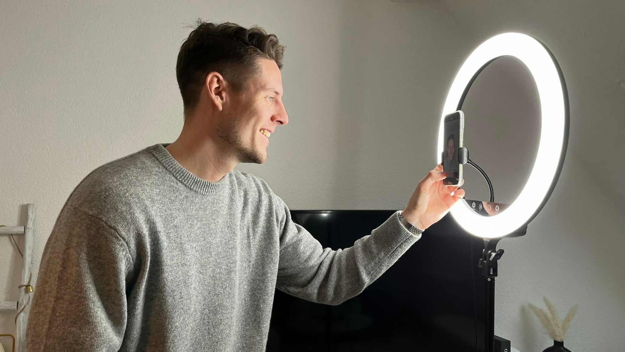 Ein Mann im grauen Pulli, mit braunen Haaren und Dreitagebart steht vor einem Ringlicht und arbeitet an seinem Smartphone.
