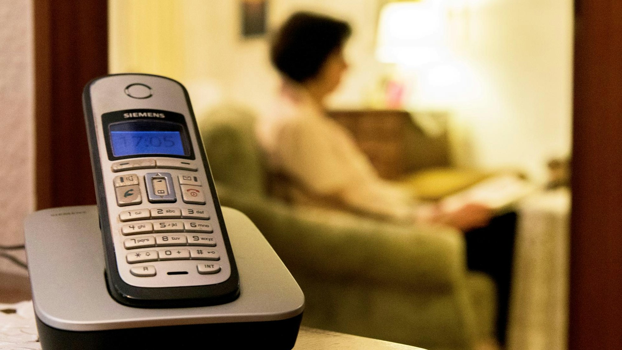 Ein Telefon, im Hintergrund sitzt eine Person im Sessel.