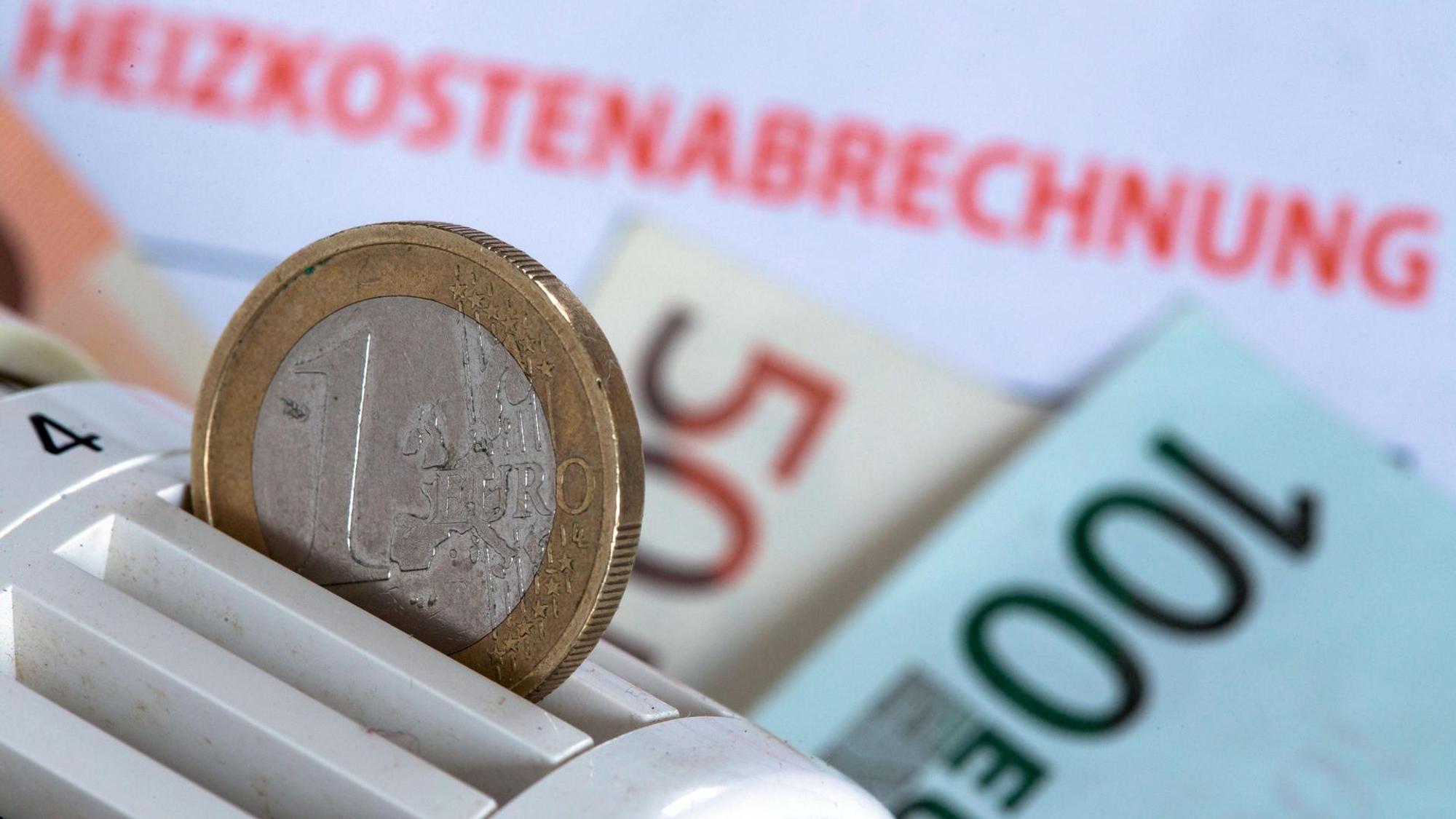 Eine Münze steckt in einem Heizungsthermostat, das auf einer Heizkostenabrechnung und Euro-Geldscheinen liegt.
