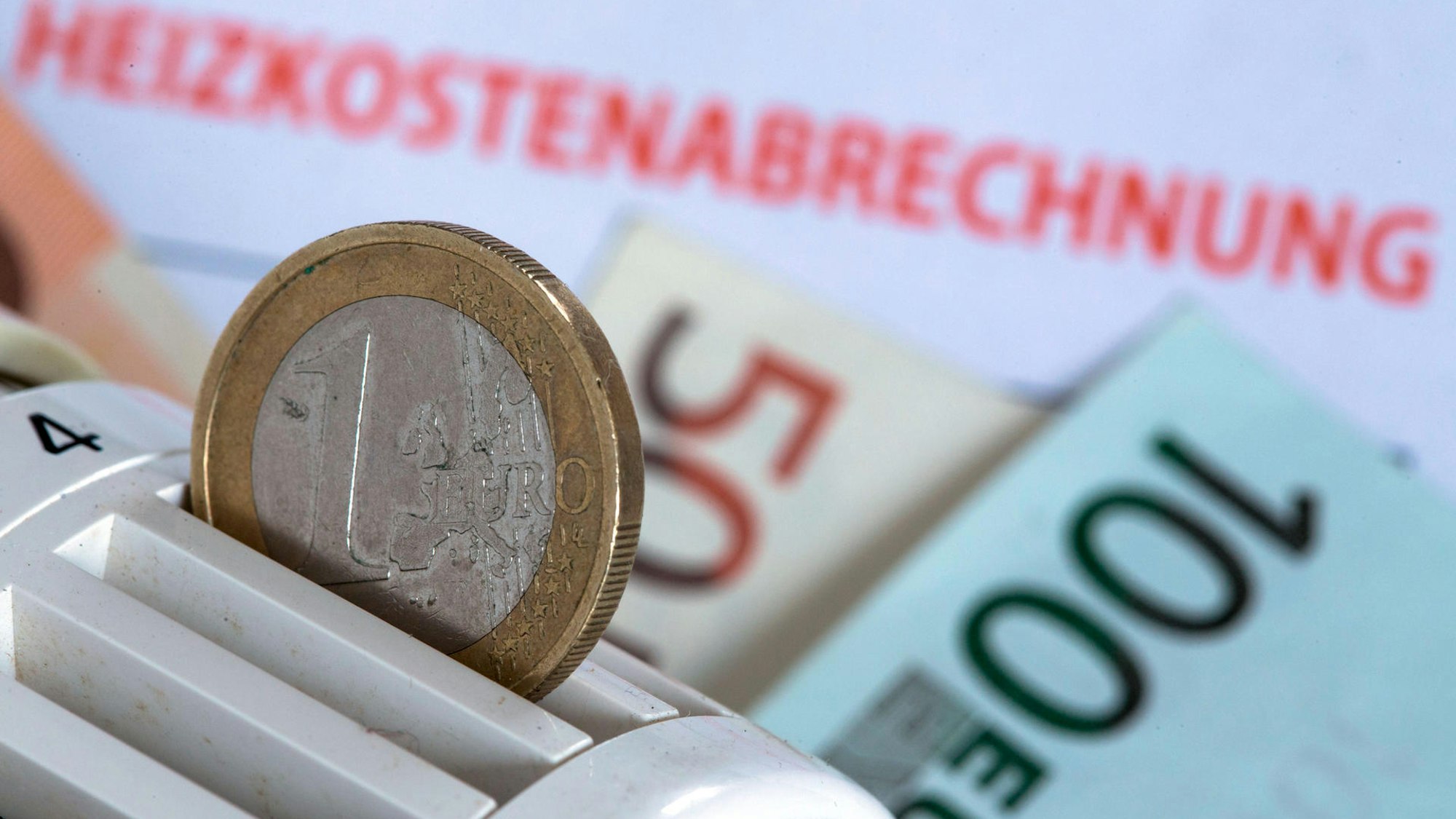 Eine Münze steckt in einem Heizungsthermostat, das auf einer Heizkostenabrechnung und Euro-Geldscheinen liegt.
