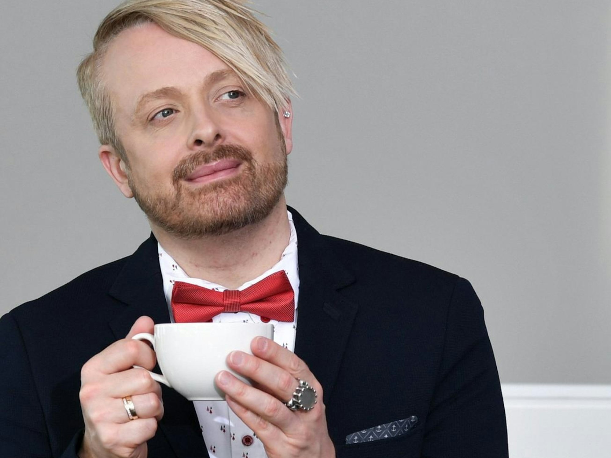 Ross Antony mit Anzug und roter Schleife. Er hält eine weiße Teetasse.