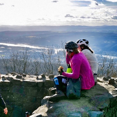 Drei Frauen machen auf dem Gipfel eines Berges Pause und sitzen dabei auf einer Steinmauer.