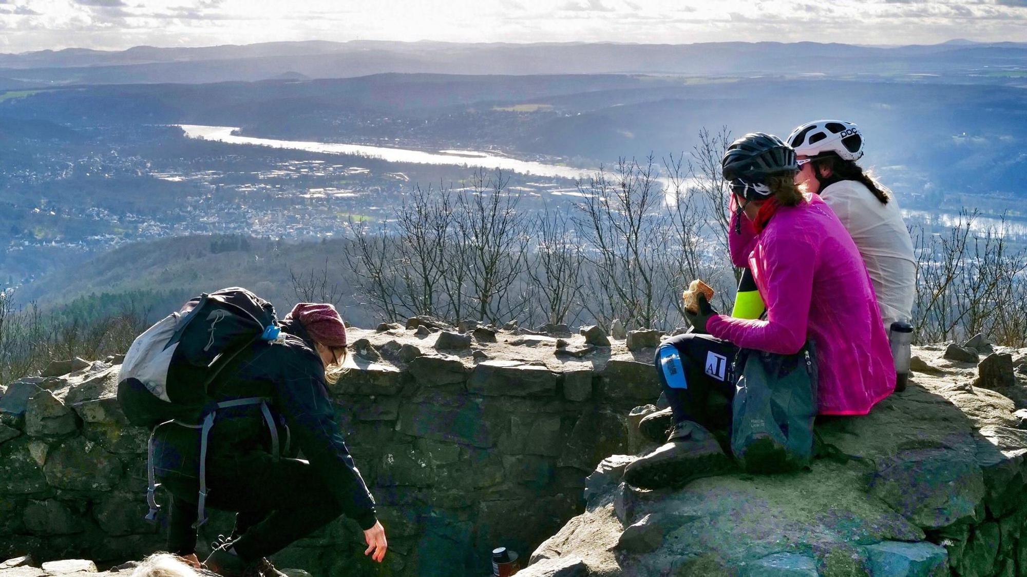 Drei Frauen machen auf dem Gipfel eines Berges Pause und sitzen dabei auf einer Steinmauer.