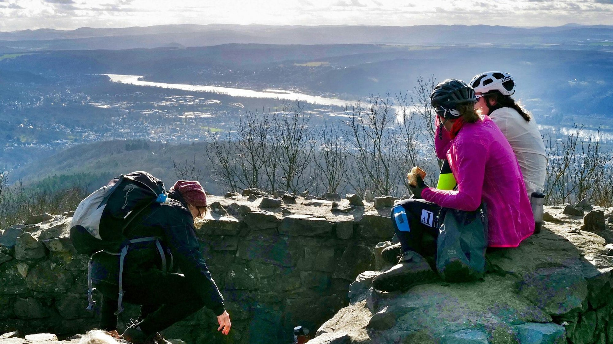 Drei Frauen machen auf dem Gipfel eines Berges Pause und sitzen dabei auf einer Steinmauer.