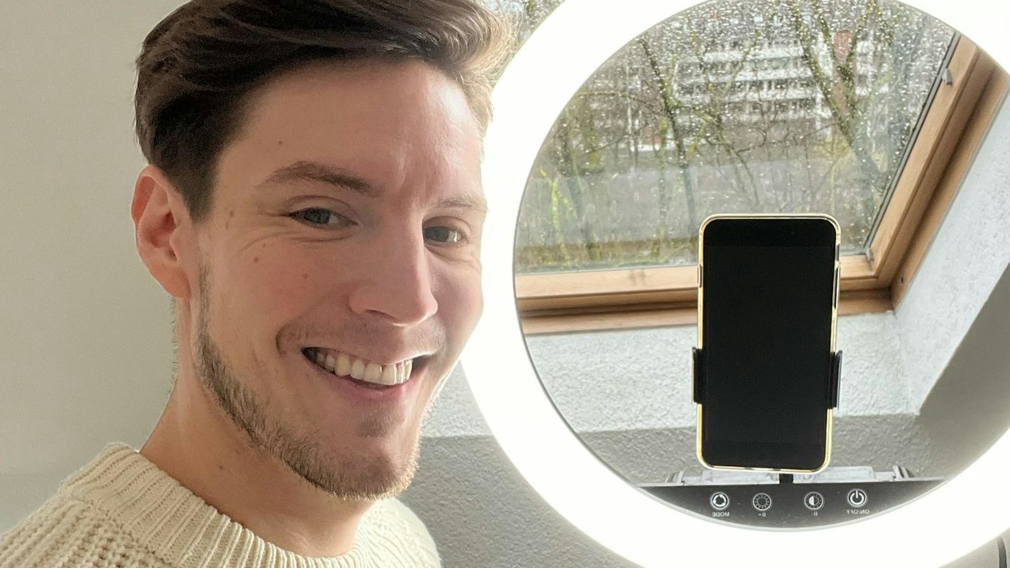 Ein Mann im grauen Pulli, mit braunen Haaren und Dreitagebart steht vor einem Ringlicht mit Smartphone. Er lacht in die Kamera