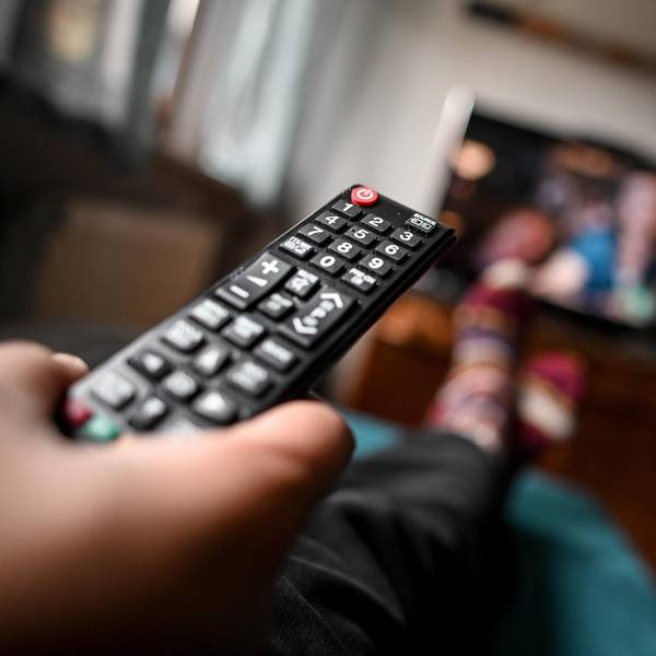 Eine Person hält eine Fernbedienung in der Hand, im Hintergrund ist unscharf ein Fernsehgerät zu sehen.