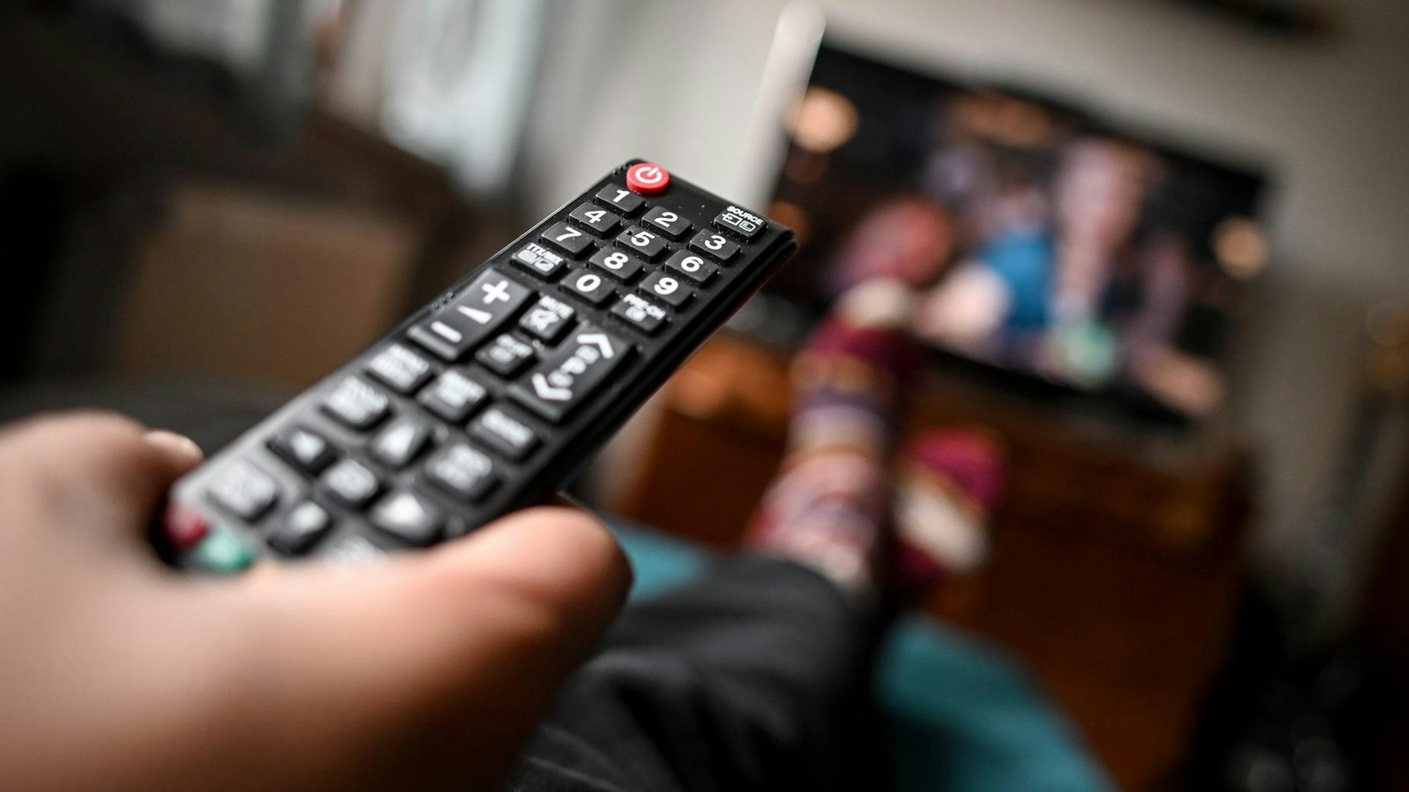 Eine Person hält eine Fernbedienung in der Hand, im Hintergrund ist unscharf ein Fernsehgerät zu sehen.