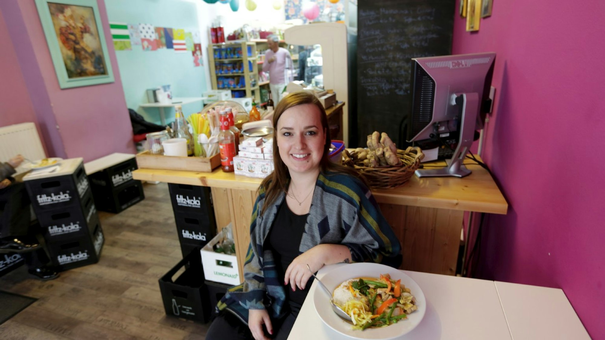 Frau sitzt an einem weißen Tisch, auf dem ein Teller mit Essen steht, im Hintergrund die Theke eines kleinen Ladens