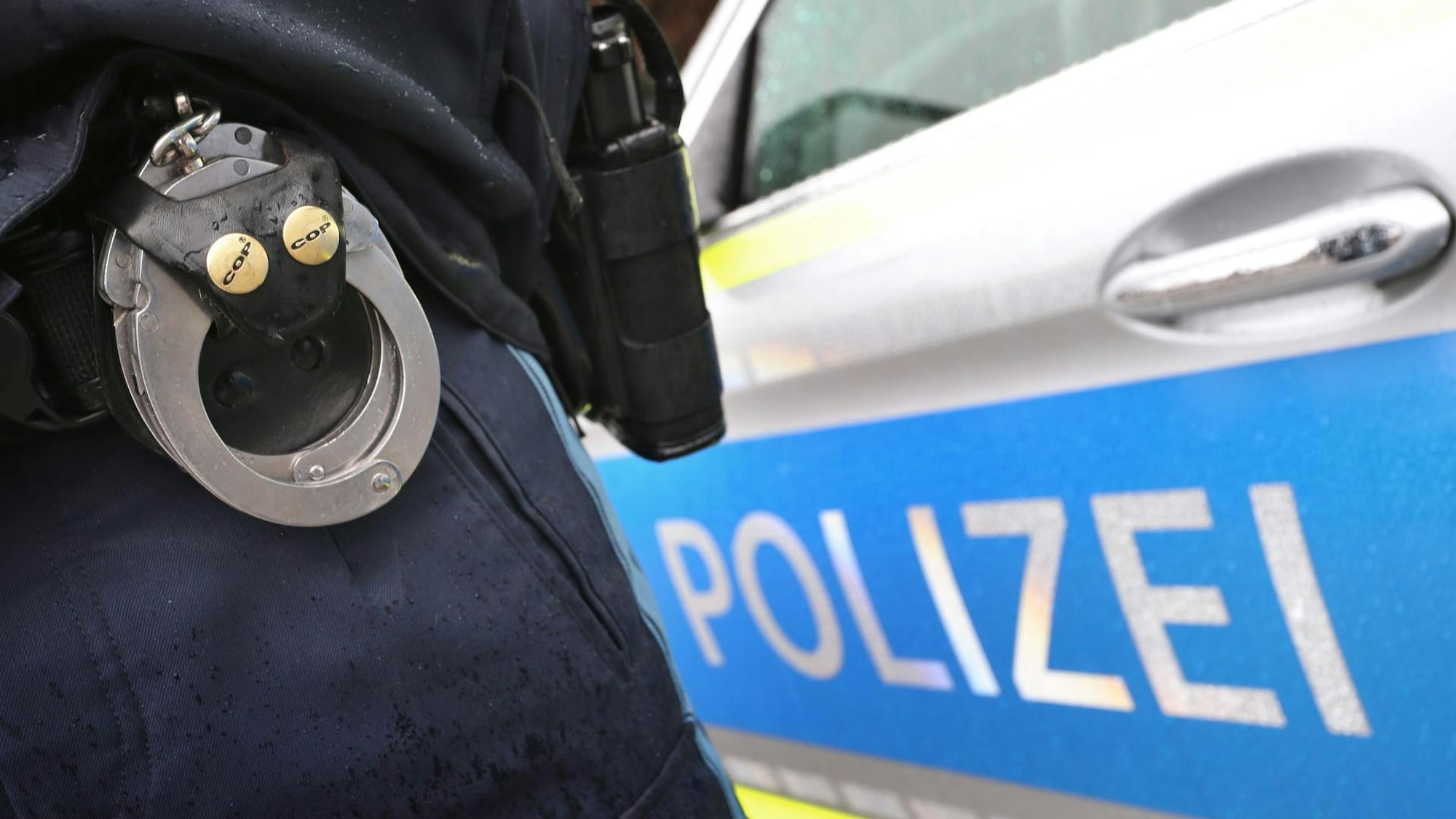 Auf dem Bild sind ein Polizeiauto und ein Polizist mit Handschellen zu sehen.