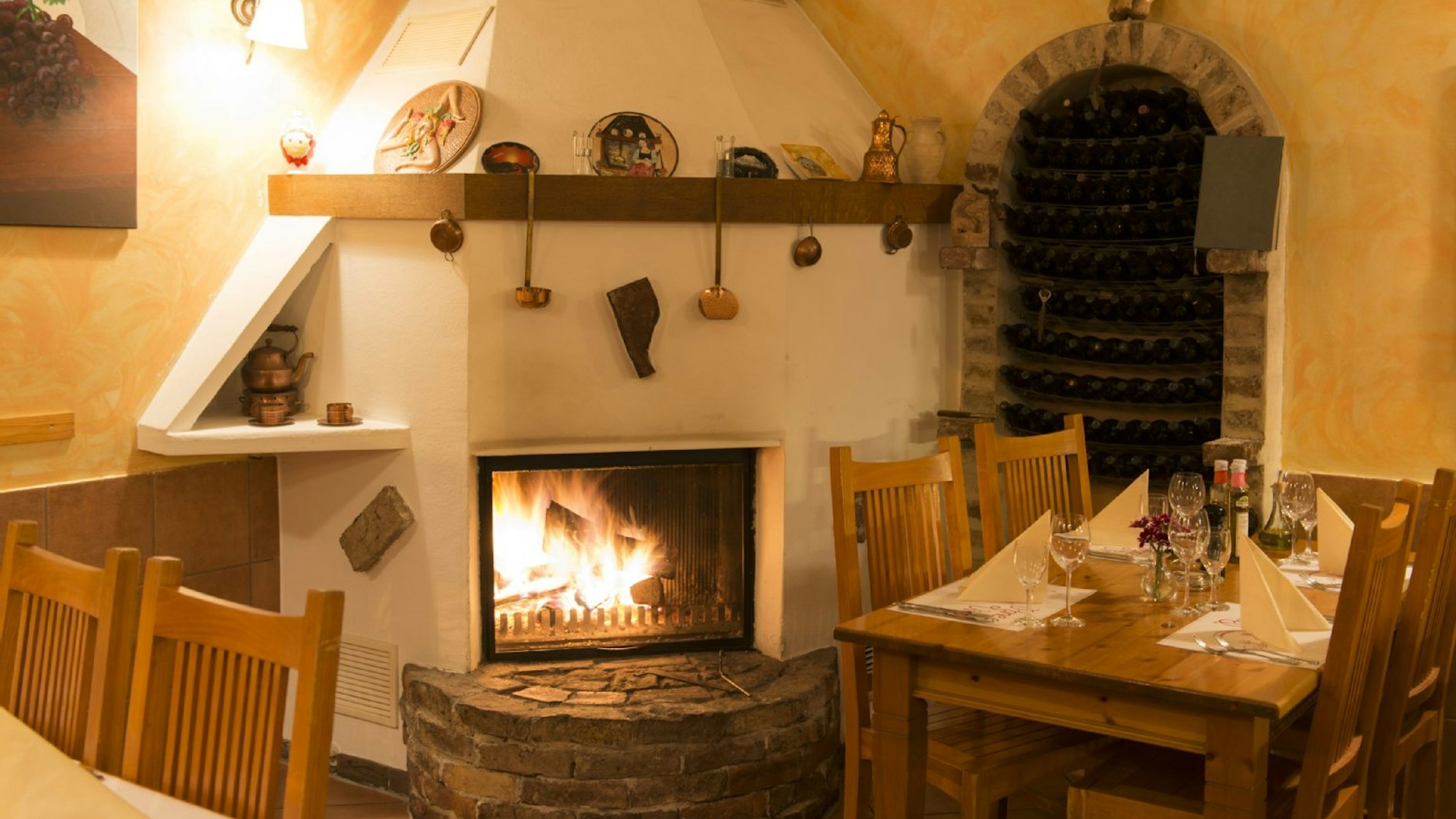 Ein rustikaler Kamin mit Steinvorbau zwischen Holztischen, geschmückt mit Suppenkellen und Tontellern im Pane e Vino in Köln.
