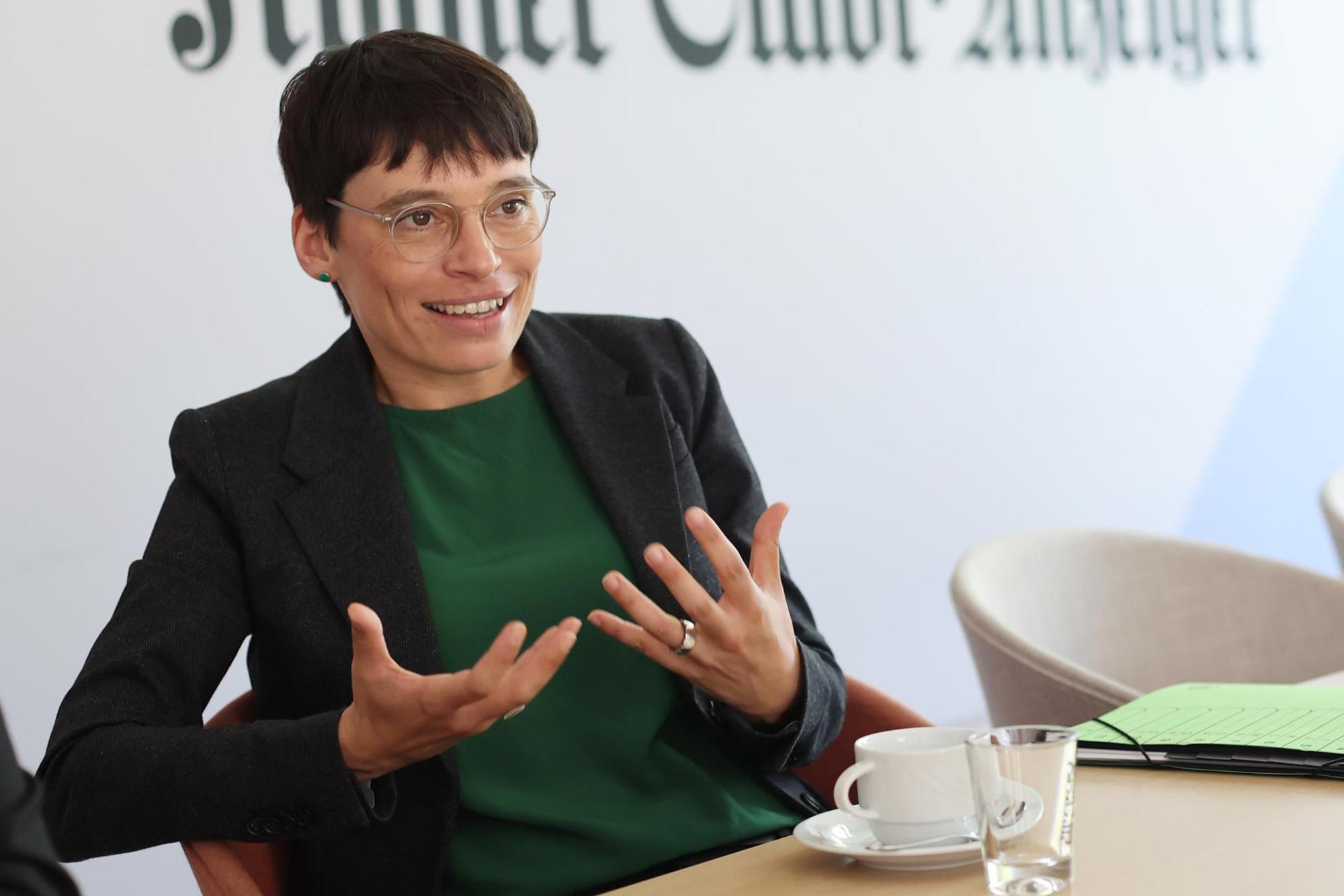 Familienministerin Josefine Paul gestikuliert mit ihren Händen. Sie trägt einen schwarzen Blazer mit einer grünen Bluse. Auf dem Tisch steht eine Teetasse und ein Glas.