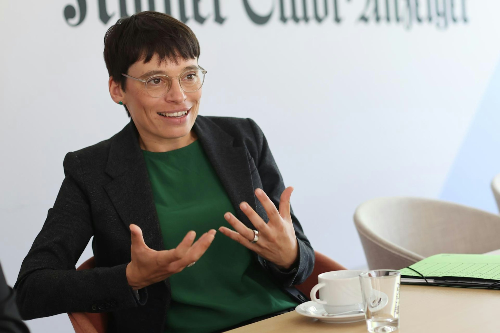 Familienministerin Josefine Paul gestikuliert mit ihren Händen. Sie trägt einen schwarzen Blazer mit einer grünen Bluse. Auf dem Tisch steht eine Teetasse und ein Glas.