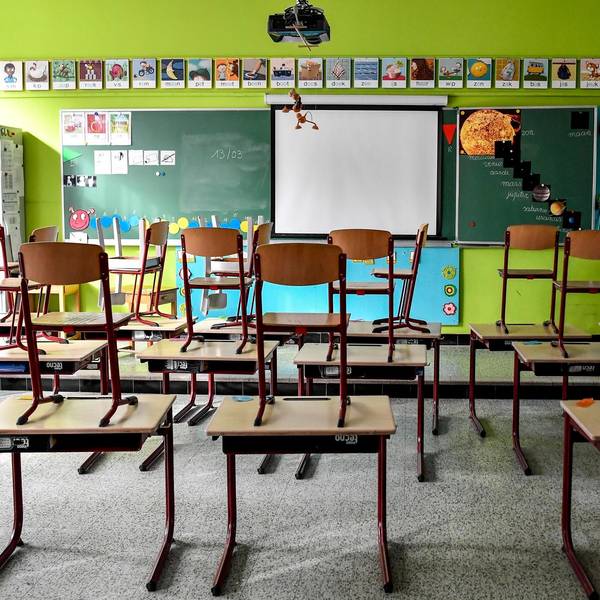In einem leeren Klassenzimmer stehen Stühle auf Tischen. Vorne ist eine Tafel zu sehen, vor der Tafel ist eine Leinwand.