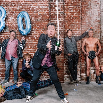 Die Prinzen feiern 30 Jahre Karriere. Der Leadsänger hält eine Champagnerflasche in der Hand, aus der Champagner in die Höhe schießt.