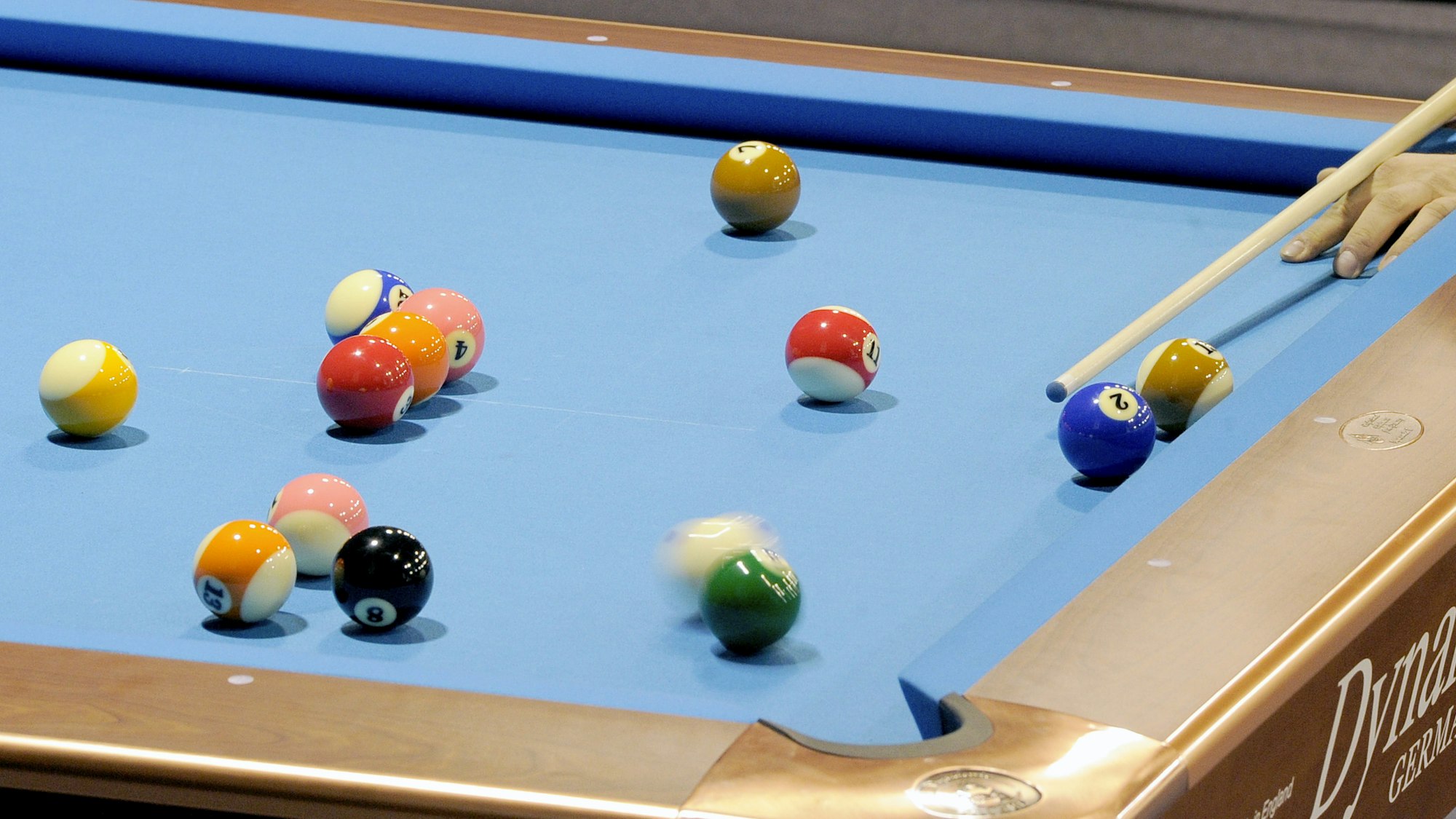 Ein Billardtisch mit vielen Kugeln. Eine Hand hält einen Kö vor die weiße Kugel.