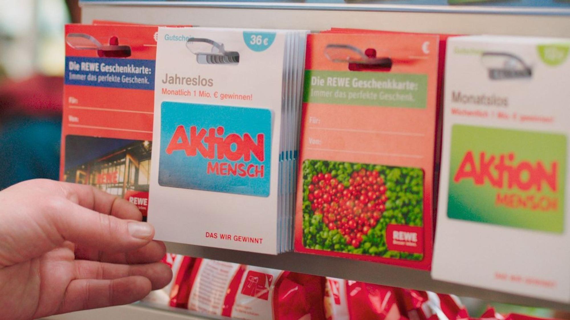 Ein Los der Aktion Mensch wird von einer Frauenhand aus einem Regal bei Rewe genommen. Daneben befinden sich weitere Lose.
