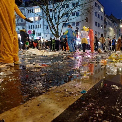 Müll liegt am späten Abend auf der Zülpicher Straße, im Hintergrund stehen kostümierte Menschen in kleinen Gruppen zusammen.