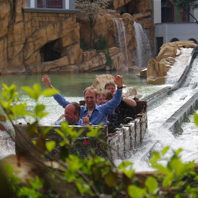 Besucher sitzen in einem Fahrgeschäft im Phantasialand, das durch eine Wasser-Fläche fährt. Ein Mann hat die Arme gehoben.