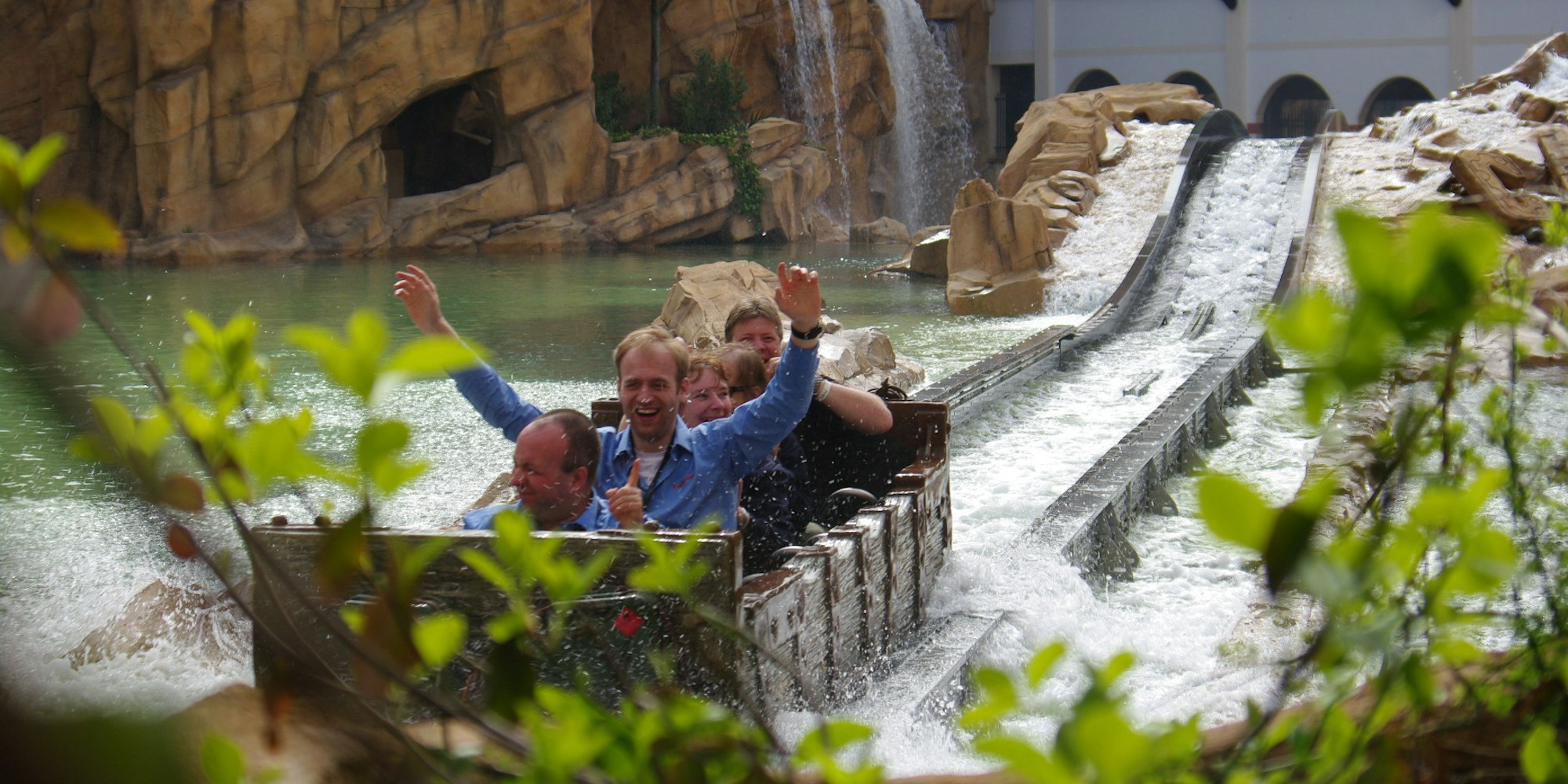 Besucher sitzen in einem Fahrgeschäft im Phantasialand, das durch eine Wasser-Fläche fährt. Ein Mann hat die Arme gehoben.