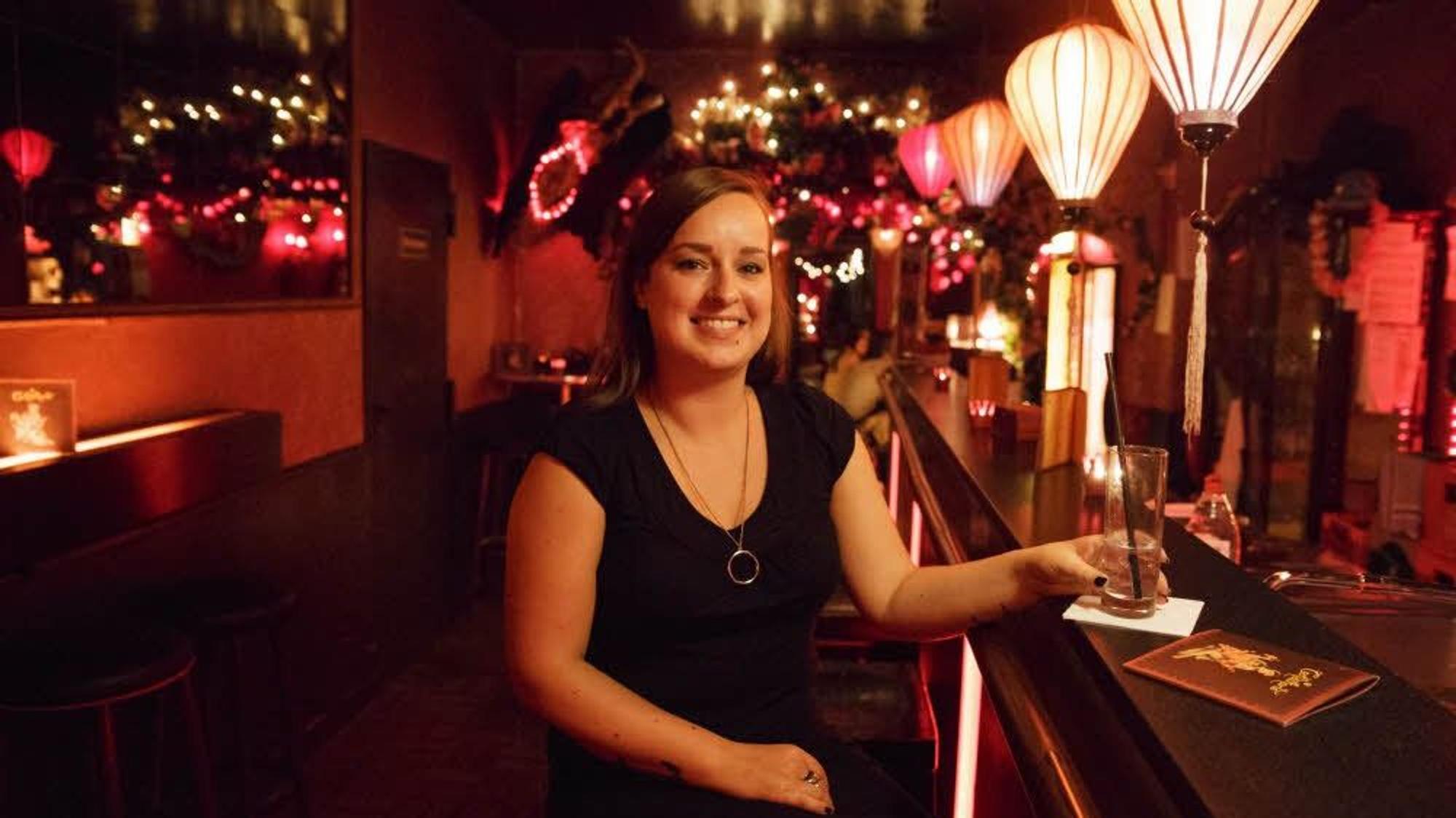 Frau sitzt mit einem Getränk in einer dunklen Bar, über ihr leuchtende Lampions