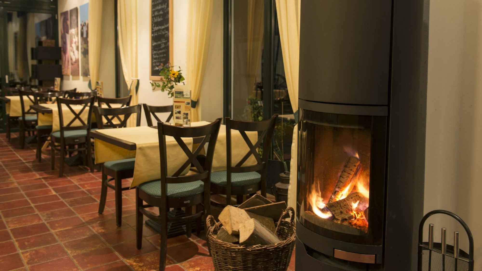 Ein schwarzer, moderner Kamin brennt in einem Raum mit Holzstühlen an Tischen mit weißen Tischdecken, der Boden ist mit Tonfliesen gedeckt.