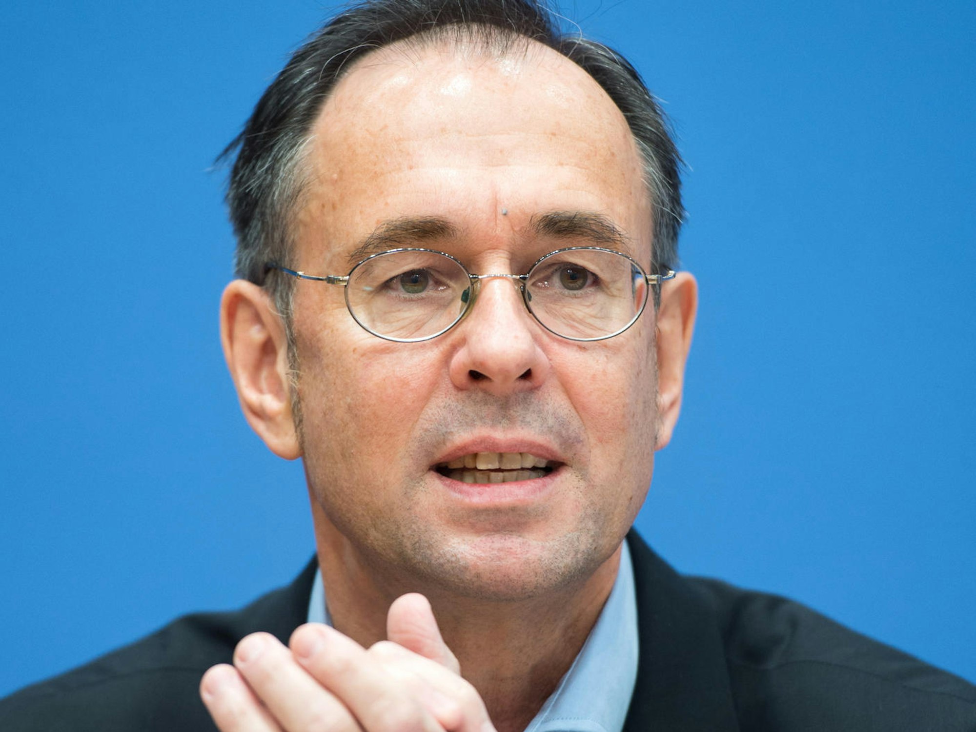 Zick guckt bei einer Pressekonferenz in die Kamera. Er trägt einen Anzug und eine Brille, der Hintergrund ist blau.