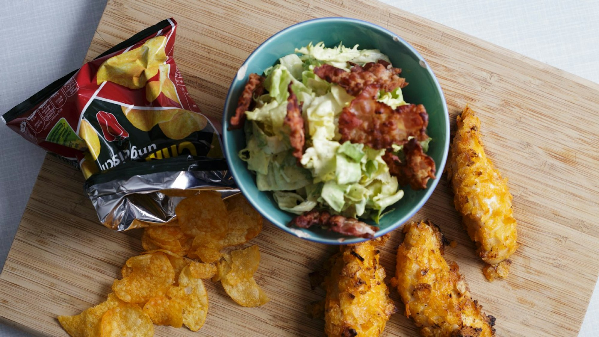 Knusprige Hähnchen-Stücke liegen auf einem Brett, daneben eine Schüssel mit Salat und eine Tüte Chips