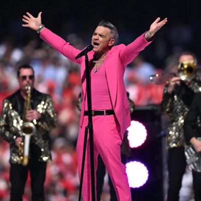 Robbie Williams streckt beim Live-Auftritt die Arme aus. Er trägt einen pinken Anzug und singt. Hinter ihm spielt seine Band mit Glitzer-Jacketts.