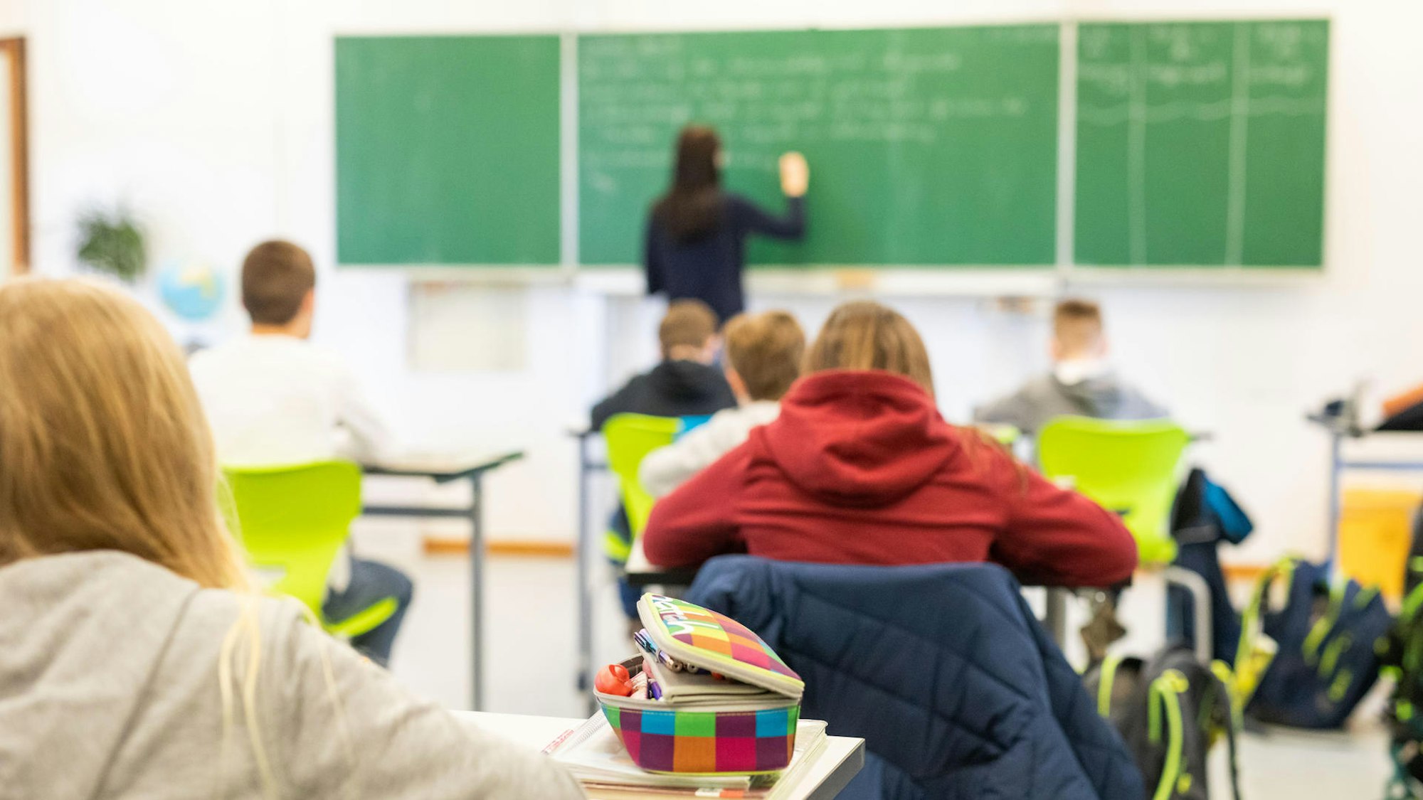 Kinder schauen im Klassenraum nach vorne an die Tafel, wo eine Lehrerin steht.
