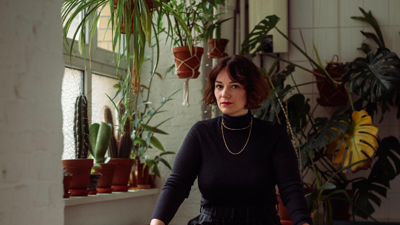 Mareice Kaiser im schwarzen Rollkragen-Pillover vor einer Wand mit Zimmerpflanzen.
