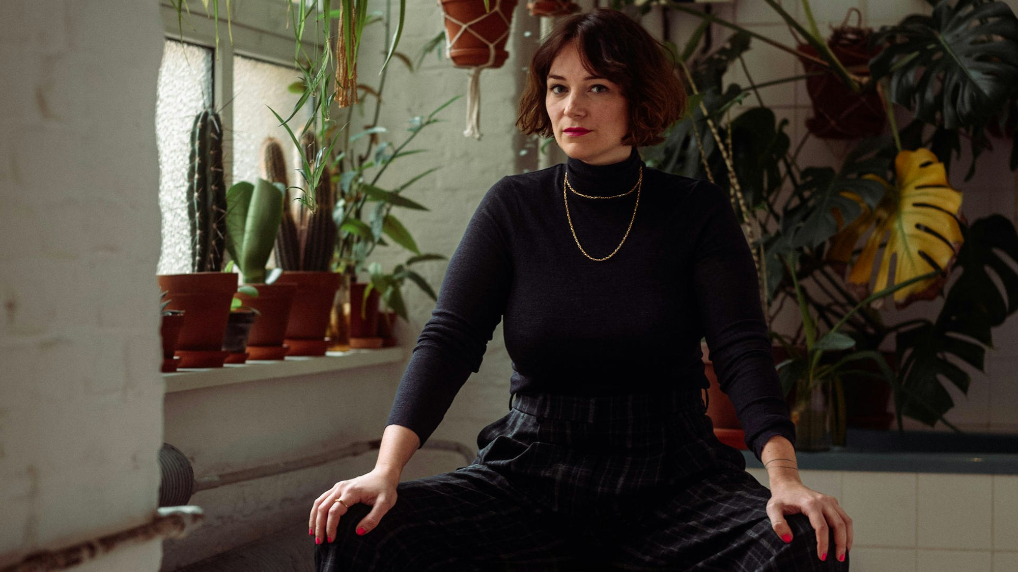 Mareice Kaiser im schwarzen Rollkragen-Pillover vor einer Wand mit Zimmerpflanzen.