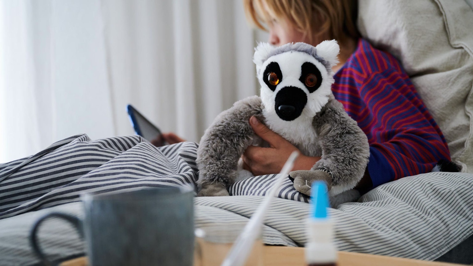 Ein Fieberthermometer, Nasenspray und eine Tasse Tee stehen vor einem Bett, in dem ein krankes Kind mit einem Kuscheltier sitzt und auf ein Tablet schaut.